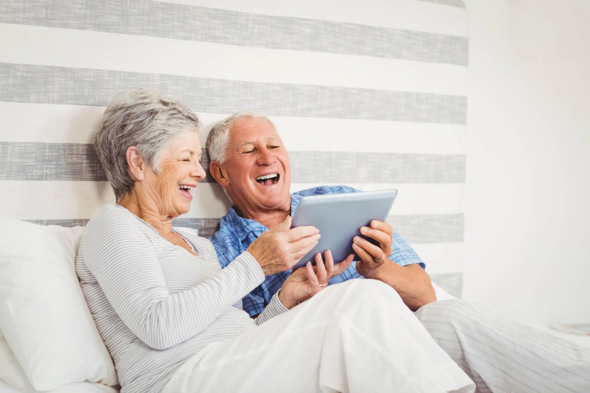 Zwei ältere Menschen, eine weibliche, eine männliche Person, gucken auf ein Tablet und lachen herzlich